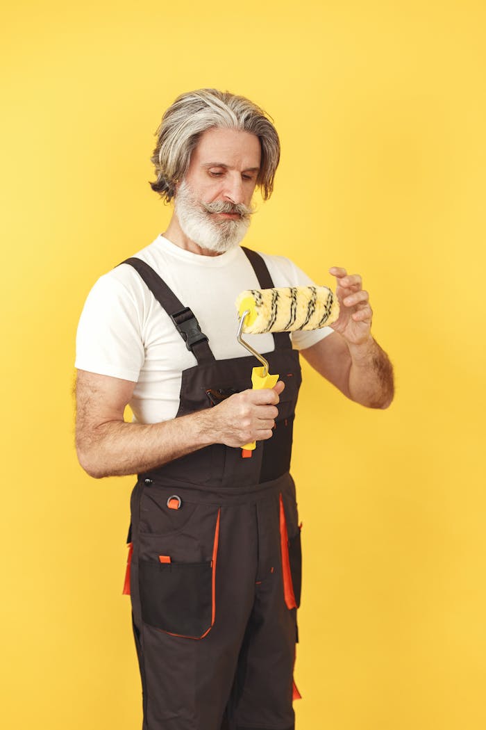 Senior man with a beard holding a paint roller against a yellow background, wearing work attire.